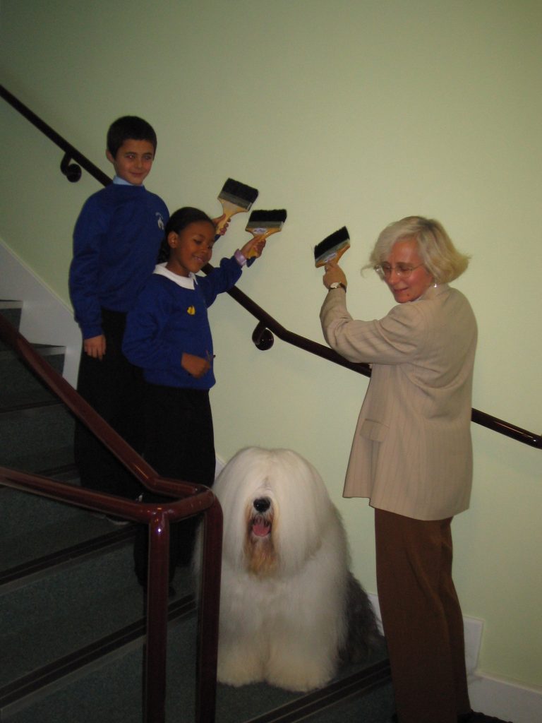A school teacher and school pupils redecorate a school. They pose next to the Dulux Dog (an Old English Sheepdog), holding paintbrushes and painting a wall in emulsion paint.