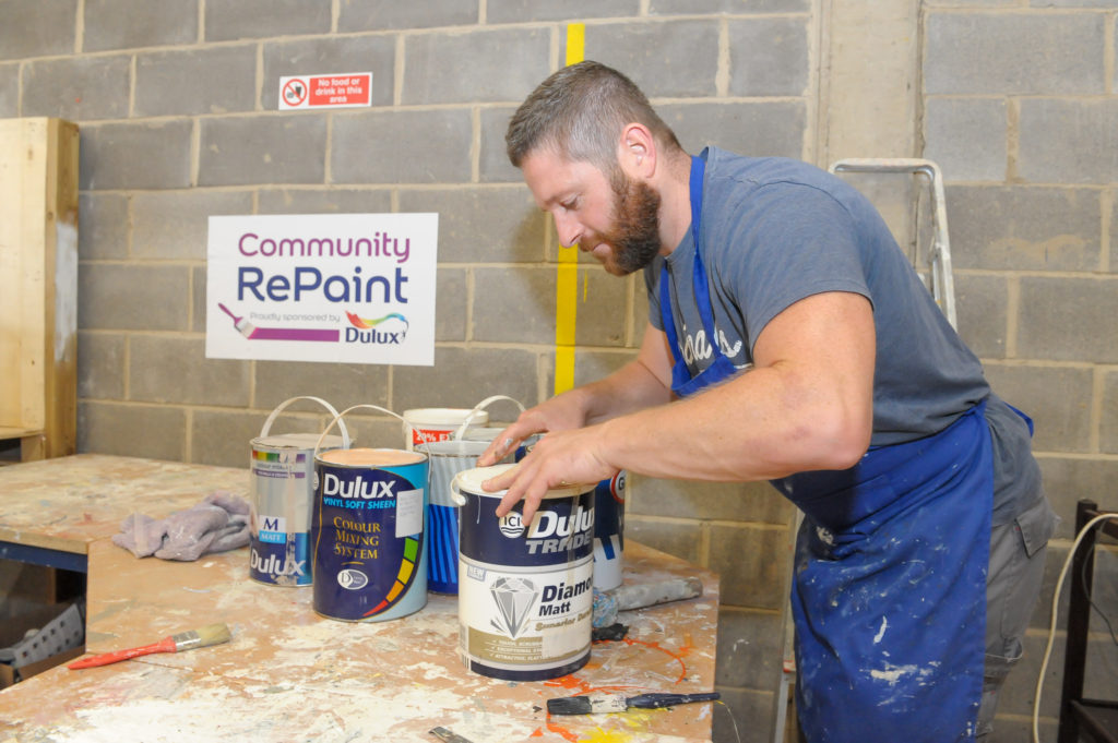 A member of staff at the Community RePaint Loughborough scheme in a workshop processing paint ready for resale. He is in the process of returning a lid to a container of Dulux Trader Diamond Matt.
