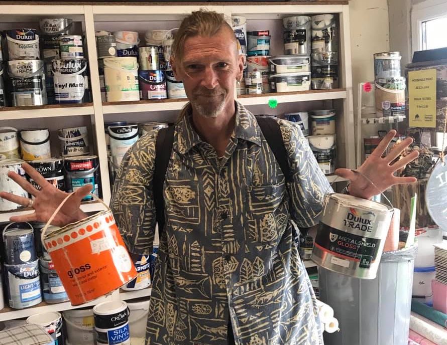 A customer poses with two cheap and recycled containers of paint purchased from Community RePaint Cornwall. In the background is a selection of reusable paint for sale.
