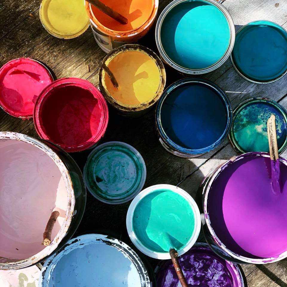 Looking into a variety of paint containers with their lids removed. There is a selection of colours including, pink paint, yellow paint, blue paint, turquoise paint, purple paint and orange paint.