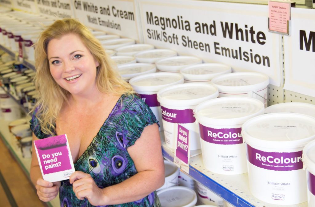 A photo of a staff member at Community RePaint Cambridgeshire holding a flyer that reads "do you need paint?". Behind are two long shelves full of low cost and eco friendly ReColour in silk emulsion, soft sheen emulsion and matt emulsion in off white, white and cream.